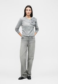 Γυναίκα που φοράει γκρι κουμπωτό ζακέτα με διακοσμητικά λουλούδια, φαρδιά ανοιχτόγριμα τζιν και μαύρα παπούτσια, στέκεται με τα χέρια πίσω από την πλάτη.