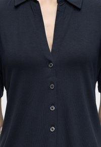 Chemise noire à manches courtes avec col et quatre boutons, portée par une personne, visible des épaules jusqu'au milieu du torse.