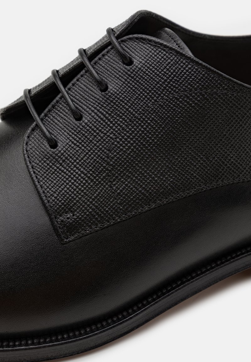 Chaussure en cuir noir avec un panneau supérieur texturé, six œillets pour les lacets, un bout rond et une finition lisse le long de la semelle.