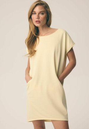 Kobieta o długich blond włosach, ubrana w luźną, krótką bladożółtą sukienkę, ręce w kieszeniach, na neutralnym tle.
