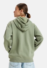 Green hooded sweatshirt with a relaxed fit, ribbed cuffs and hem, and soft texture. Styled with light blue denim jeans.