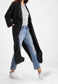 Lungo cappotto nero con un design drappeggiato, indossato sopra una blusa a righe, abbinato a jeans strappati azzurri e sneakers bianche.