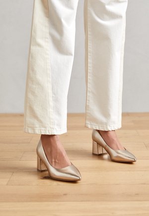 Metalliske guld højhælede sko med spids tå og en kraftig gennemsigtig blokhæll. Båret med hvide breddebukser på et trægulv.