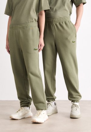 Pantalones de chándal verdes con cintura elástica, bolsillos laterales y logo de Lacoste. Combinados con zapatillas de color claro, aptos para uso casual.