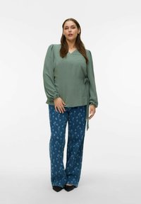 Grønn innpakket topp med lange ermer og knutedetalj, kombinert med blå vide jeans med blomsterprint. Glatt stofftekstur.