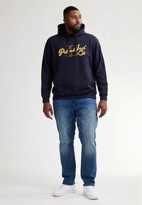 Felpa con cappuccio blu navy con testo grafico giallo, materiale in misto cotone, tasca a marsupio frontale, abbinata a jeans blu sbiaditi e sneakers bianche.