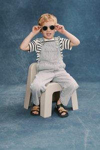 Ριγέ μπλουζάκι με κοντά μανίκια; ανοιχτό μπλε σαλοπέτα με λεπτές λευκές ρίγες; μπεζ πλαστική καρέκλα; μαύρα και πράσινα σανδάλια; στρογγυλά γυαλιά ηλίου.