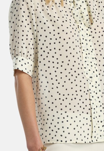 Enyhén texturált krémszínű blúz szórt fekete pöttyös mintával, rövid, fodros ujjakkal és félgombos lezárással.