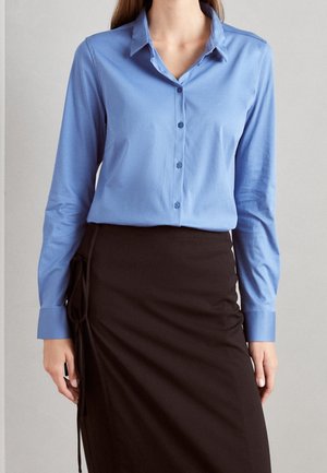 Kobieta ubrana w jasnoniebieską, zapinaną na guziki koszulę z długim rękawem, wsuniętą w wysoką czarną spódnicę z wiązaniem po bokach.