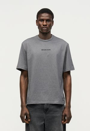 Mann trägt ein graues Michael Kors T-Shirt mit schwarzem Logo-Schriftzug, kombiniert mit dunkelgrauen Jeans, stehend vor einem einfarbigen hellen Hintergrund.