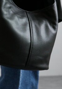 Musta nahkainen käsilaukku, jossa on pehmeä rakenne, saumaton ja pyöreä muoto, viimeistelty hienovaraisilla ompeluioneilla.