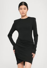 Jonge vrouw met zwart haar in een strakke bun, gekleed in een aansluitende zwarte jurk met lange mouwen, gerimpelde details en een asymmetrische zoom.