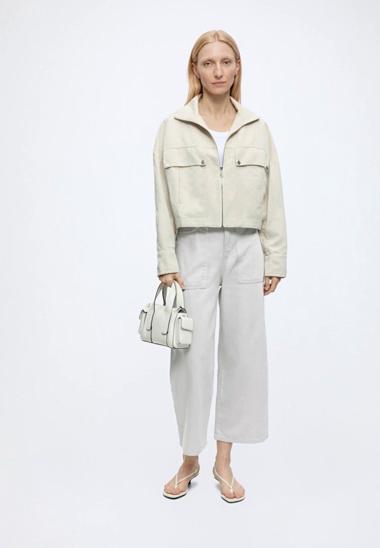 Femme debout face à l'avant, portant une veste crème, un pantalon blanc à jambes larges, des sandales blanches et tenant un petit sac à main crème. Fond uni.