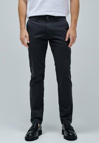 Pantaloni in cotone neri, slim-fit, con una texture liscia, dotati di chiusura con bottone e passanti per cintura, abbinati a scarpe nere senza lacci.