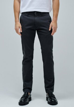 Pantalons noirs en coton slim-fit avec une texture lisse, dotés d'une fermeture à bouton et de passants de ceinture, associés à des chaussures noires à enfiler.