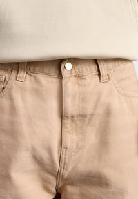 Pantalones de algodón beige con corte recto, que presentan un botón metálico dorado y bolsillos delanteros, mostrando una superficie de tela lisa y texturizada.