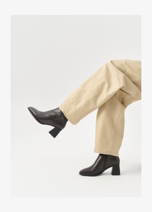 Botines de cuero negro hasta el tobillo con punta afilada y un tacón grueso, combinados con pantalones de pierna ancha beige. Diseño sencillo, textura suave.