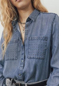 Persona che indossa una camicia di jeans blu con borchie metalliche sulle tasche del petto e collane d'argento con un ciondolo a forma di lucchetto.