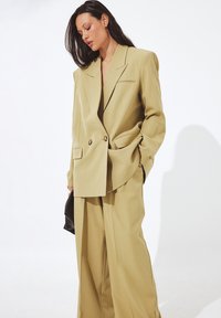 Divpusējais khaki bleizeris ar polsterētām plecu daļām un asiem atlokiem, savienots ar platām biksēm, stilizēts ar melnu somu.