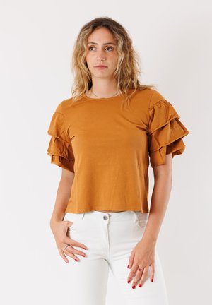 Top corto color ruggine con dettagli a volant sovrapposti sulle maniche, realizzato in un materiale morbido, abbinato a pantaloni bianchi aderenti.