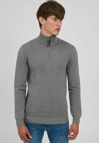 Suéter gris con cuello de cremallera que presenta un patrón texturizado en zigzag, puños y dobladillo acanalados, y un diseño de corte slim. Combinado con jeans de mezclilla azul.