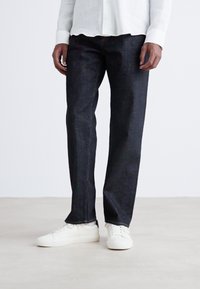Jeans de mezclilla oscura con corte recto, que presentan un diseño clásico, costuras sutiles y se combinan con zapatillas blancas.