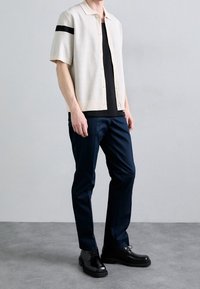 Polo a maniche corte in maglia color avorio con un motivo testurizzato e una striscia nera sulla spalla sinistra, abbinato a pantaloni blu scuro e scarpe nere.