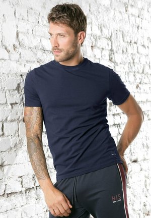 Navyblaues T-Shirt mit kurzen Ärmeln aus glattem Stoff, mit Rundhalsausschnitt. Kombiniert mit schwarzen Sporthosen. Sichtbare Tattoos am Unterarm.
