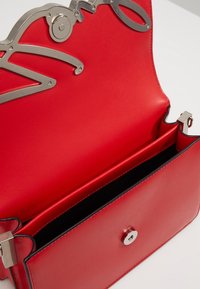 Červená kožená kabelka s otevřeným klopovým víkem, zdobená stříbrným kovovým hardwarem a magnetickým uzávěrem, umístěná na bílé ploše.