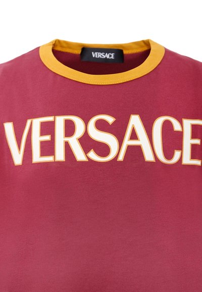 T-shirt in cotone marrone con colletto a coste giallo e la scritta "VERSACE" stampata in lettere bianche in grassetto sul petto. Texture liscia, maniche corte.
