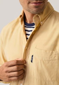Camisa de botones de pana amarilla con un bolsillo en el pecho y botones blancos, superpuesta sobre una camisa de rayas azul y blanca. Tejido texturizado.