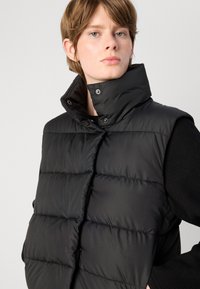 Colete acolchoado preto com gola alta e fecho de pressão. Apresenta secções acolchoadas e uma textura suave. Usado por cima de um suéter de malha preto.