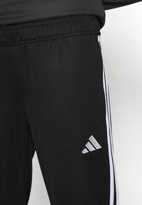 Černé sportovní kalhoty s hladkou texturou, zdobené bílými bočními pruhy a vyšitým stříbrným logem na spodní části nohavice.