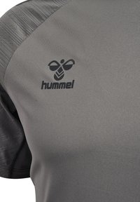Close-up van grijs sportshirtstof met zwart hummel-bijlogo en merknaam op het borstgedeelte.