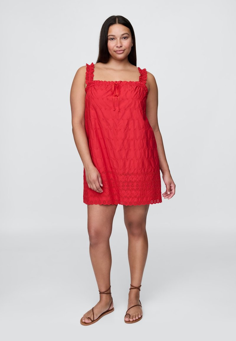 Piros ujjatlan ruha fodros pántokkal, elasztikus nyakkivágással, texturált anyagból és virágmintás lyukacsos szegéllyel. Barna pántos szandállal viselve.