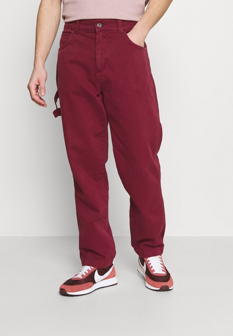 Burgunderfarbene Baumwoll-Cargohosen mit lockerer Passform, die über Taschen an den Seiten und Gürtelschlaufen verfügen. Kombiniert mit rot-weißen Turnschuhen.