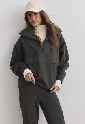 Femme portant une veste zippée gris foncé, un pantalon assorti, un col roulé crème, des gants sans doigts et une casquette de baseball crème devant un mur blanc.