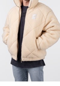 Beige quiltad fleece-jacka med dragkedja, hög krage och logotypdetalj. Texturerat material och avslappnad passform. Bärs över en mörk skjorta.