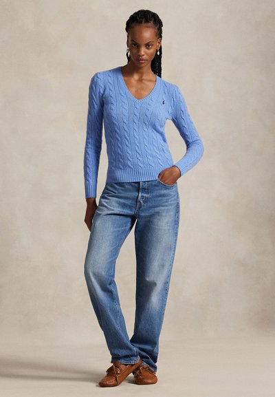 Fonott hajjal rendelkező nő, aki világoskék, fonott mintás V-nyakú pulóvert, kék farmert és barna mokaszin cipőt visel, az egyik kezét a zsebében tartva áll.