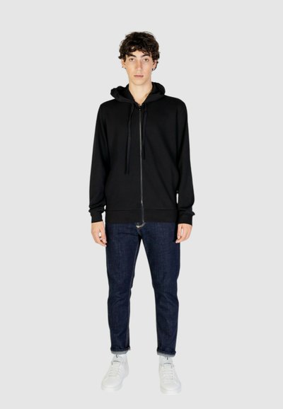 Felpa nera con zip e cappuccio regolabile, indossata sopra jeans scuri e sneakers bianche, presentata su uno sfondo neutro.