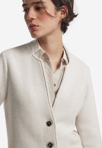 Jasno beżowy sweter z dzianiny o fakturowanym wzorze, z kołnierzem z wycięciami oraz dwoma okrągłymi czarnymi guzikami z przodu. Nałożony na jasną koszulę.
