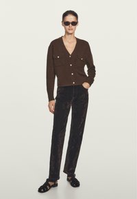 Cardigan marrone lavorato a maglia con due tasche sul petto e bottoni dorati, abbinato a pantaloni di velluto neri e sandali neri con cinturini.
