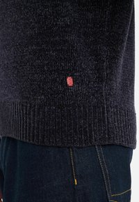 Sötét tengerészkék pulóver, texturált felülettel és bordázott szegéllyel, oldalán egy kis piros logóval, sötét farmernadrággal párosítva.