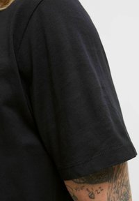 Schwarzes Kurzarm-T-Shirt aus Baumwolle mit Rundhalsausschnitt. Glatte Textur mit sichtbaren Nähten entlang des Saums und der Ärmelkanten.