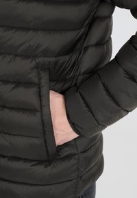 Svart puffjacka med quiltade sektioner, med en sidoficka och en närbild av en hand som vilar på fickan. Slät textur.