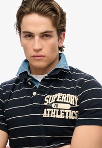 Navy gestreept polo shirt met een denim kraag. Kenmerkt zich door witte borduursels met de tekst "SUPERDRY 72 ATHLETICS." Gestructureerde stof, korte mouwen.