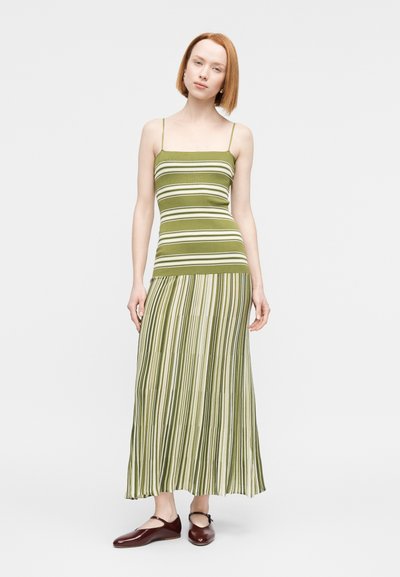 Γυναίκα με κοντά κόκκινα μαλλιά, που φοράει φόρεμα χωρίς μανίκια με πράσινες και λευκές ρίγες και σκούρα κόκκινα επίπεδα παπούτσια, στέκεται μπροστά σε ένα μονόχρωμο λευκό φόντο.