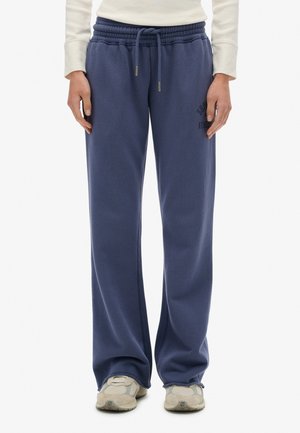 Person med løstsiddende marineblå sweatpants med elastisk talje og snor, hvide sneakers og en cremefarvet langærmet top.