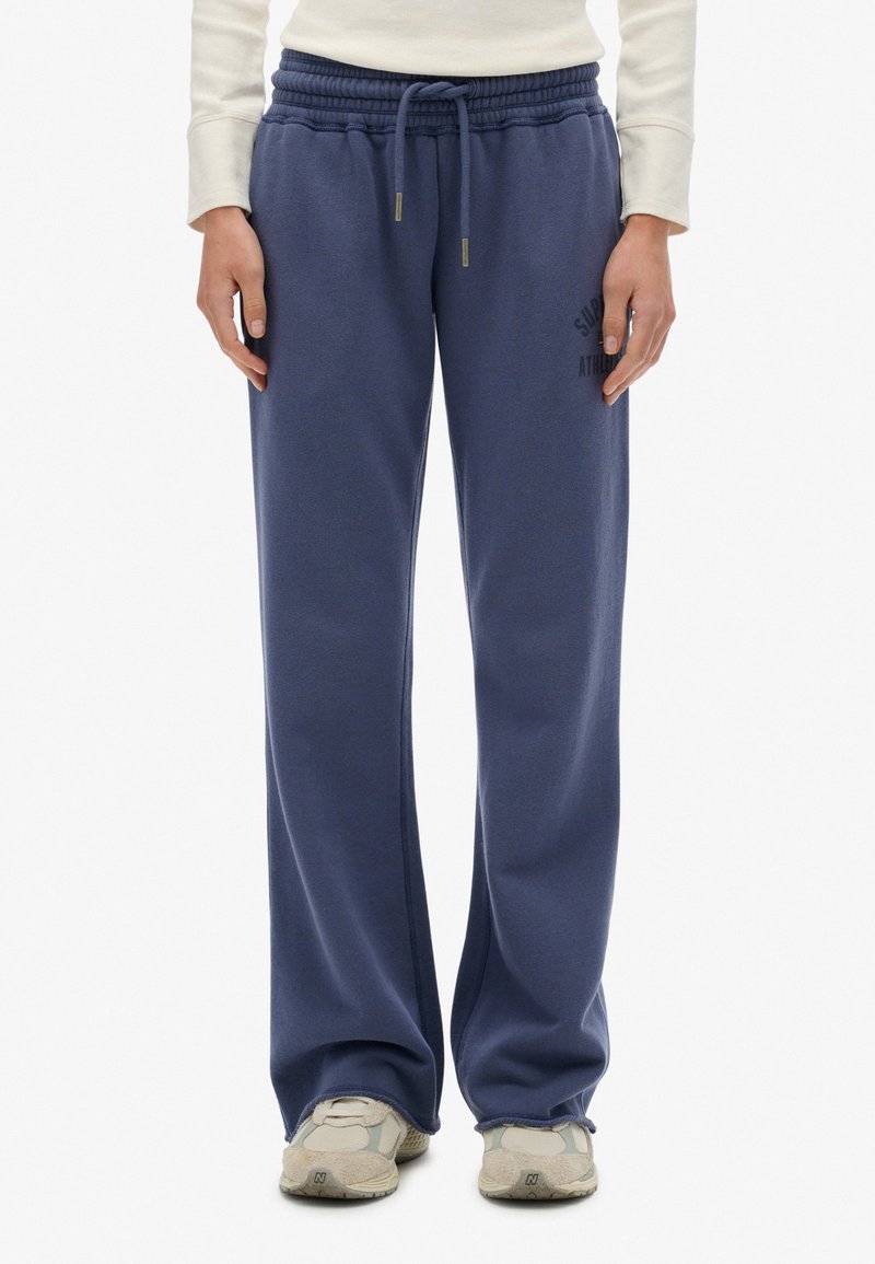 Persona que lleva puesta un pantalón de chándal azul marino holgado con cintura elástica y cordón, zapatillas deportivas blancas y una camiseta de manga larga color crema.