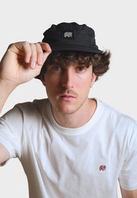 Čierna čiapka s textúrovanou látkou a štvorcovým logom s slonom. Nosí sa naklonená, v kombinácii s jednoduchým bielym tričkom.